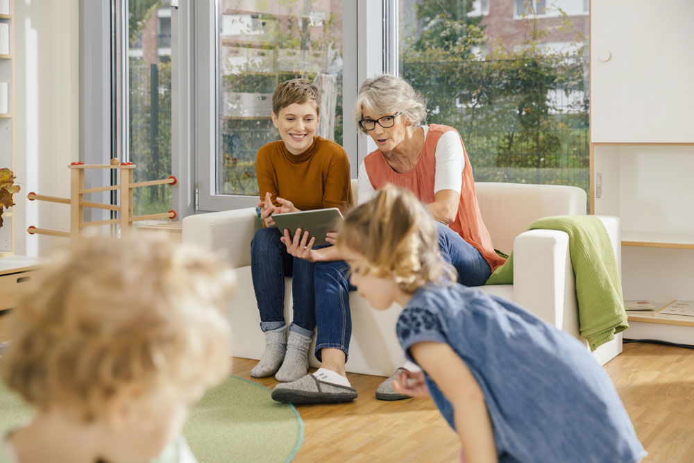 Foto zeigt zwei Erzieherinnen in einer Kita. Sie schauen auf die Kinder im Vordergrund des Bildes