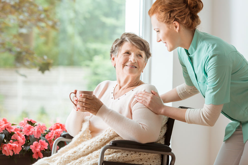 Foto zeigt alte Frau im Rollstuhl im Seniorenheim, mit helfender Pflegerin an der Seite